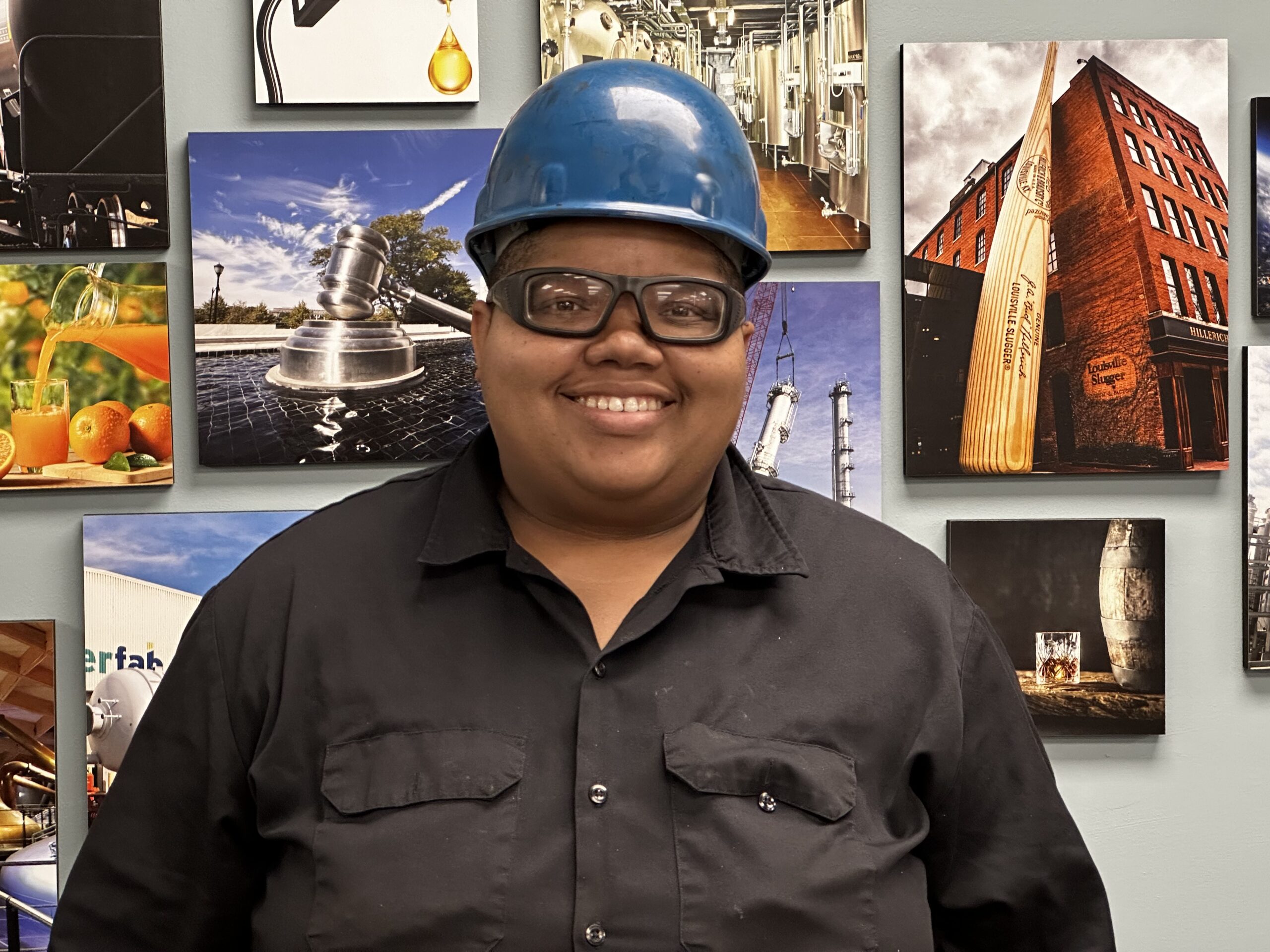 Machine operator Arie Broom stands in front of a Brighton photo collage. She's wearing a hard hat, protective eyewear, and a black button-up shirt.