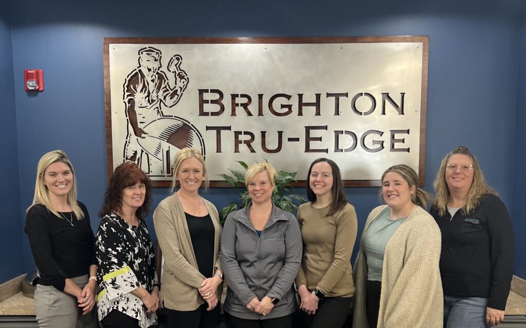 7 women employees at Brighton stand in front of the Brighton Tru-Edge sign, which rests on a dark blue wall.